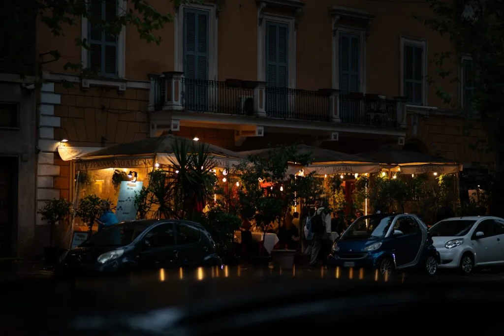 Terrasse éclairée d'une pizzeria romaine sous des auvents, voitures garées